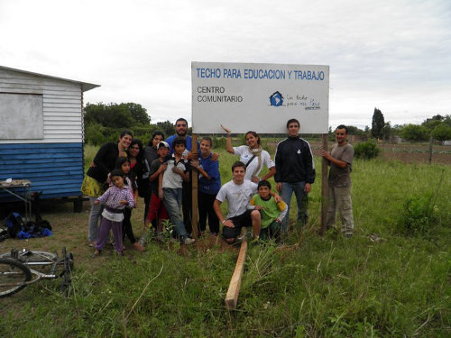 Miembros del capítulo argentino de Un techo para mi país posan frente a una valla que reza 'Techo para educación y trabajo - Centro comunitario - Un techo para mi país'
